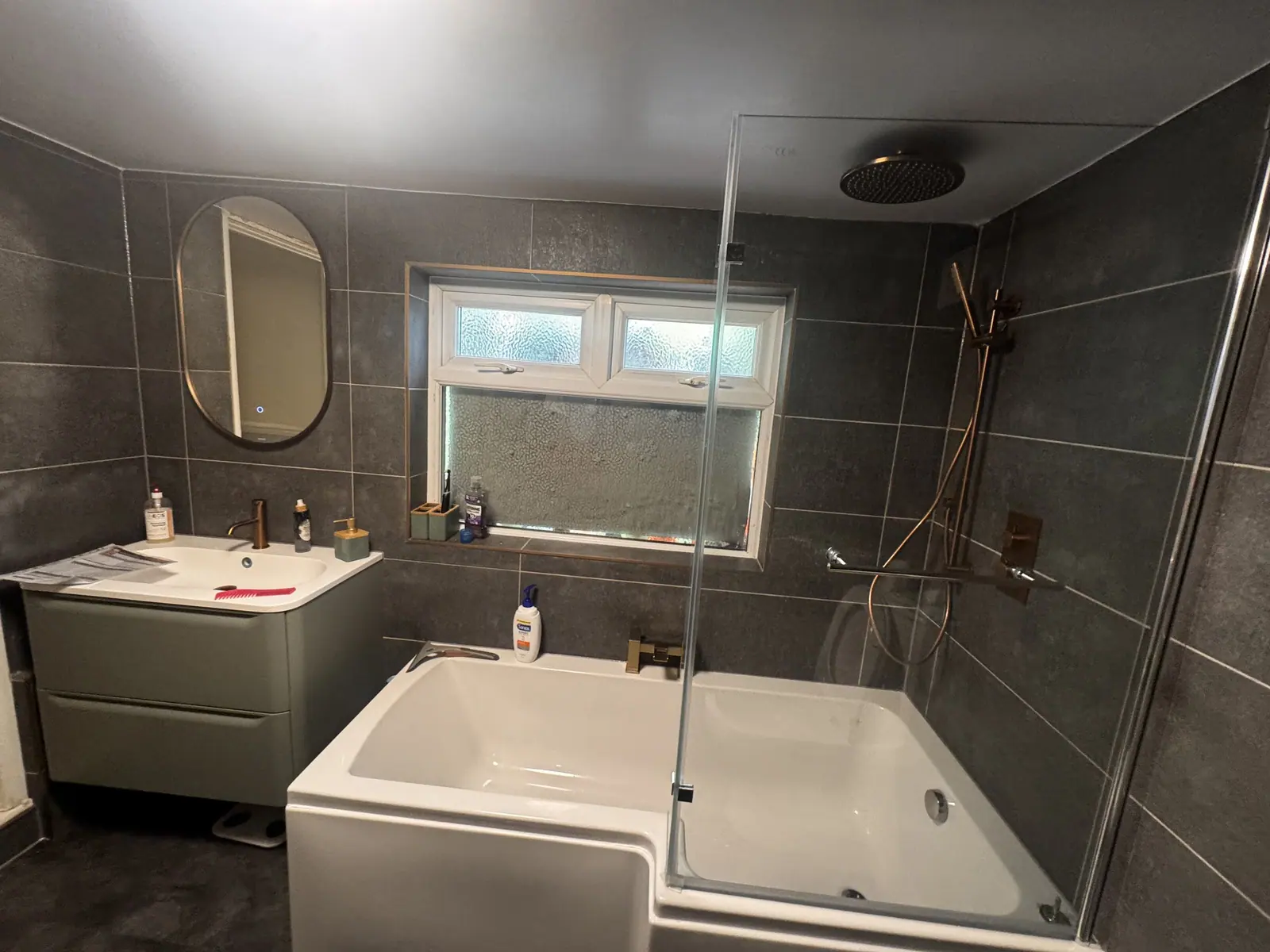 Bathroom in charcoal tiles with an L-shaped shower bath, glass screen and sage green vanity, fitted by Basingstoke Bathrooms