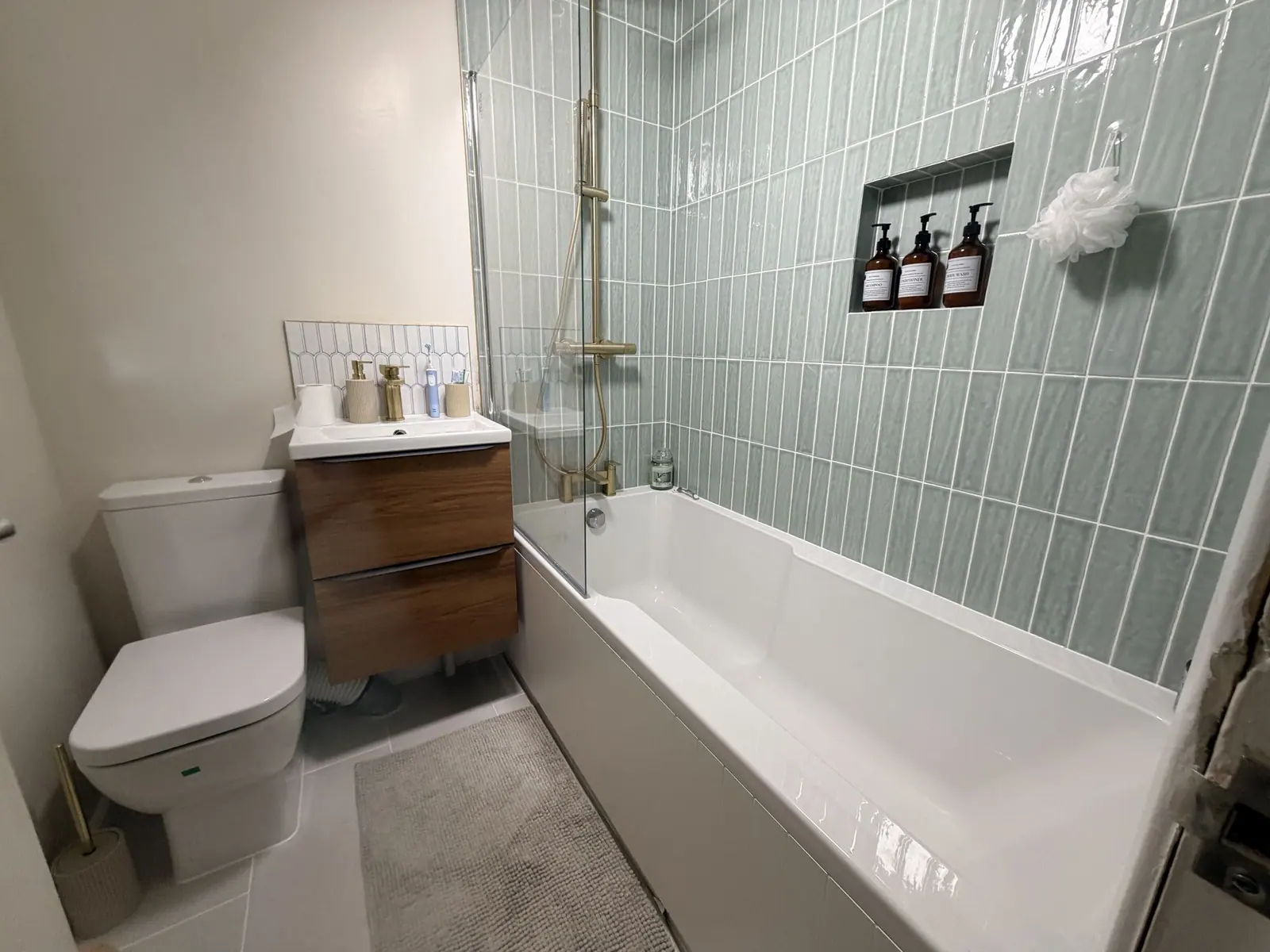 Bathroom with sage green stacked tiles, brass shower over the bath, walnut vanity and black towel rail, fitted by Basingstoke Bathrooms