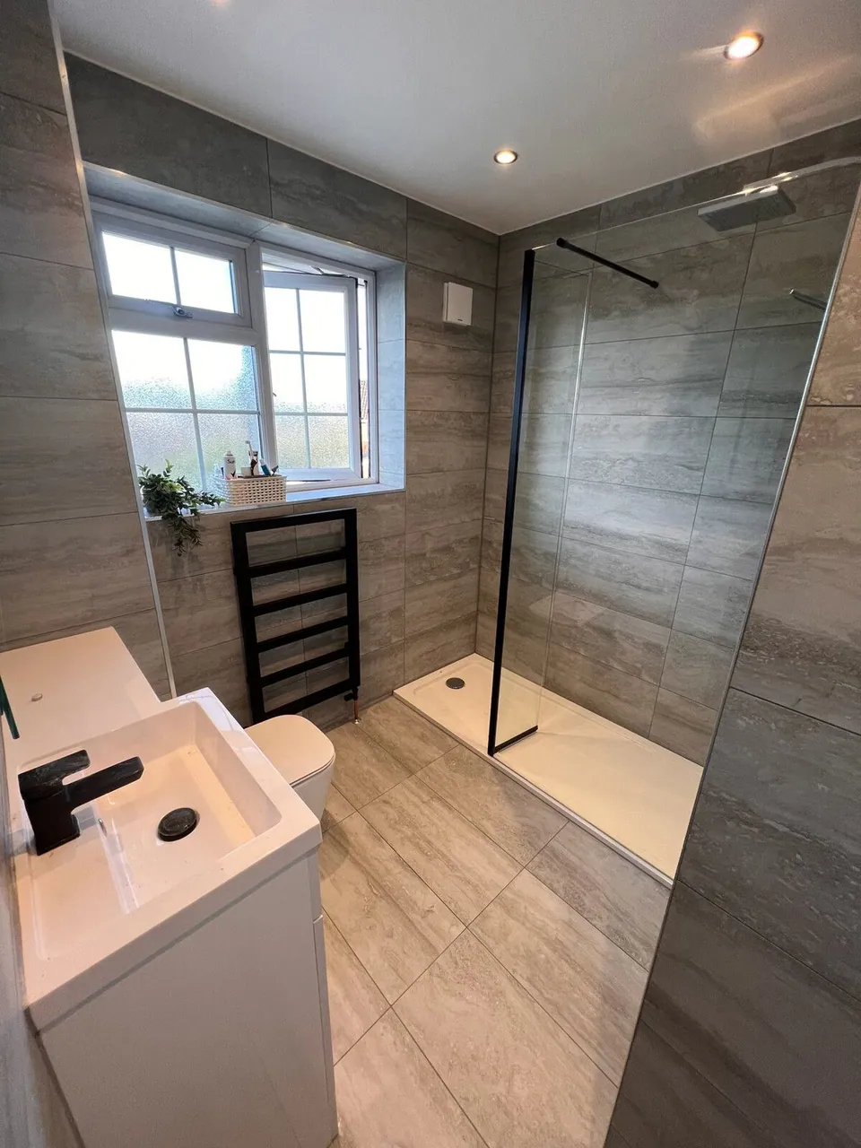 Walk-in shower with a black-framed fixed screen, grey wood-effect tiles and black basin tap, fitted by Basingstoke Bathrooms